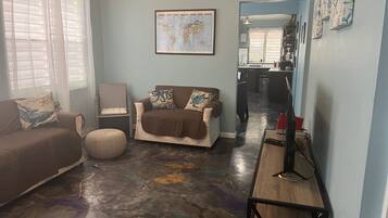 Een woonkamer met een bruine bank, een stoel en een salontafel. Aan de muur hangt een wereldkaart en er is een keuken in de achtergrond.