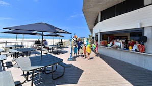 Ett utomhusområde med sittplatser, picknickbord och parasoller, med utsikt över en strand.