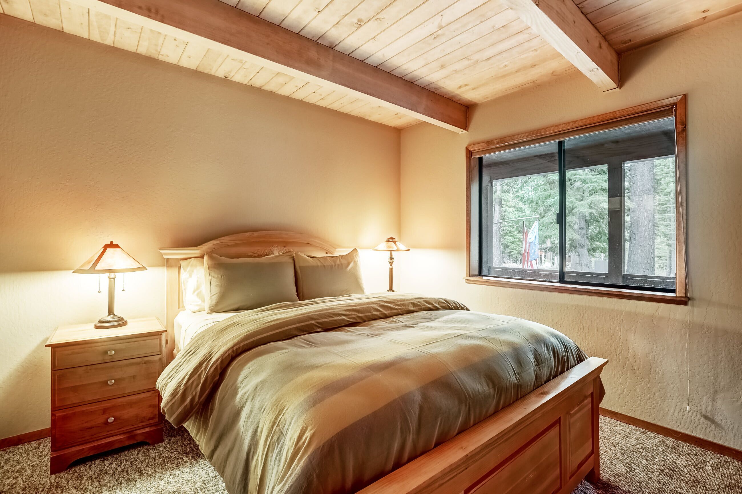 A bedroom with a wooden bed, two bedside tables with lamps, a window with a view of trees, and a sloped ceiling.