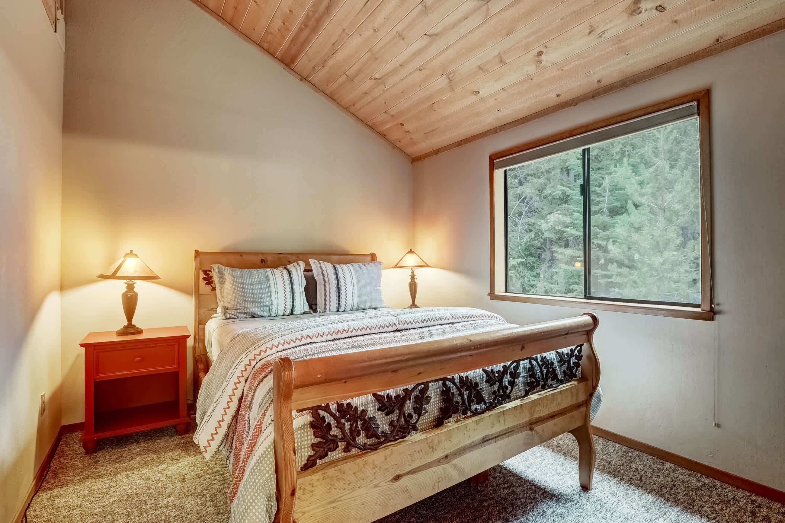 Una camera da letto con soffitto in legno, una grande finestra vista foresta, un letto con copriletto decorato e due lampade da comodino.