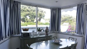 Ein Essbereich mit rundem Tisch und Stühlen, einem Fenster mit Vorhängen, einer Mikrowelle, einer Vase mit Blumen und einem Korb auf der Fensterbank.