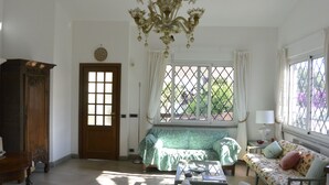 Ein Zimmer mit Holztür, einem grünen Sofa, einem Sofa mit floralem Muster, einem Glastisch und einer Chandelier.
