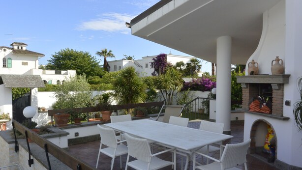 Eine Terrasse mit einem weißen Tisch und Stühlen, umgeben von Topfpflanzen und einem Backofen aus Ziegeln.