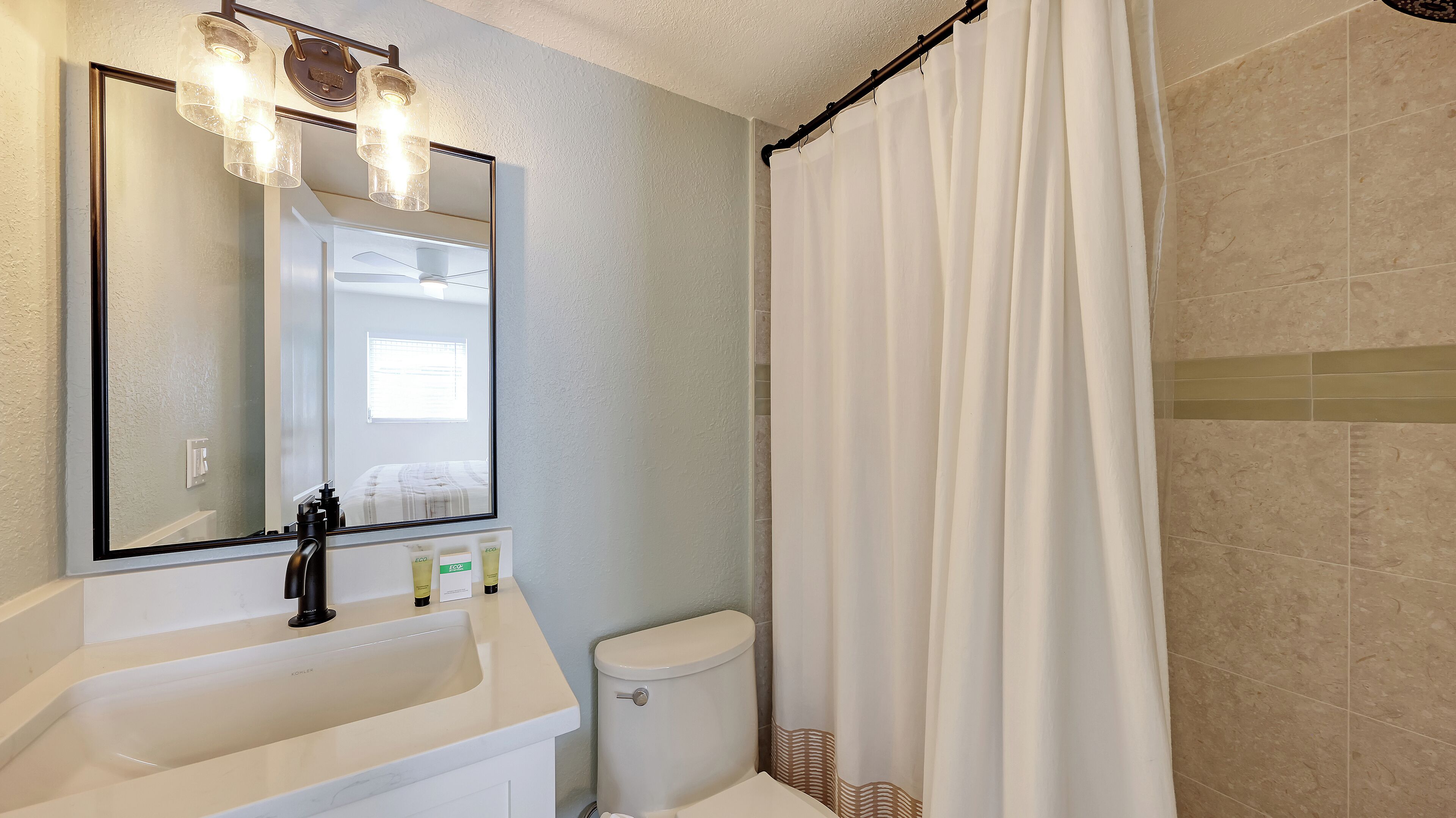 Een badkamer met een wit wastafelmeubel, toilet, douchegordijn en een spiegel met verlichting.
