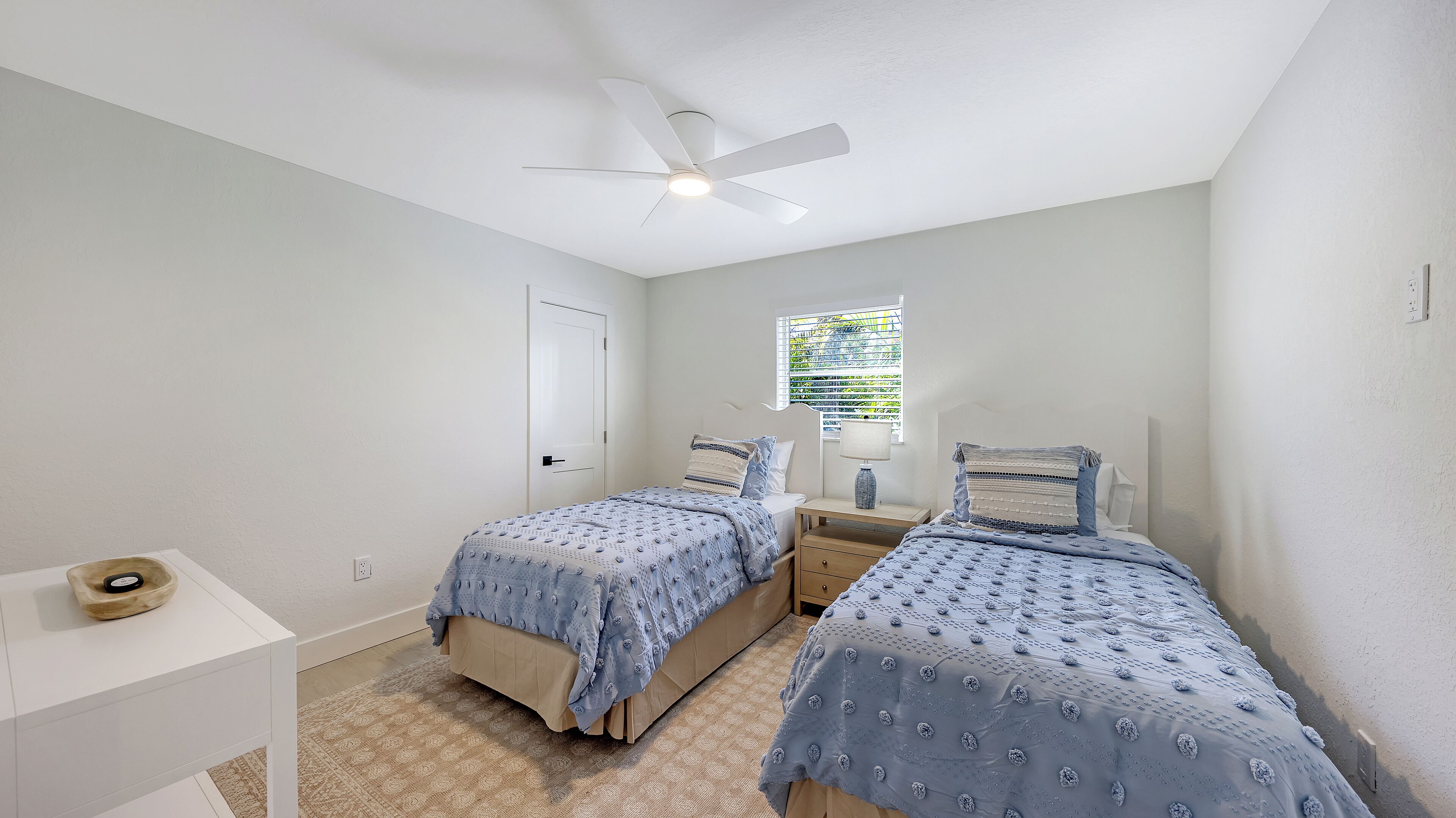 A bedroom with two beds, a ceiling fan, a window with blinds, and a small bedside table.