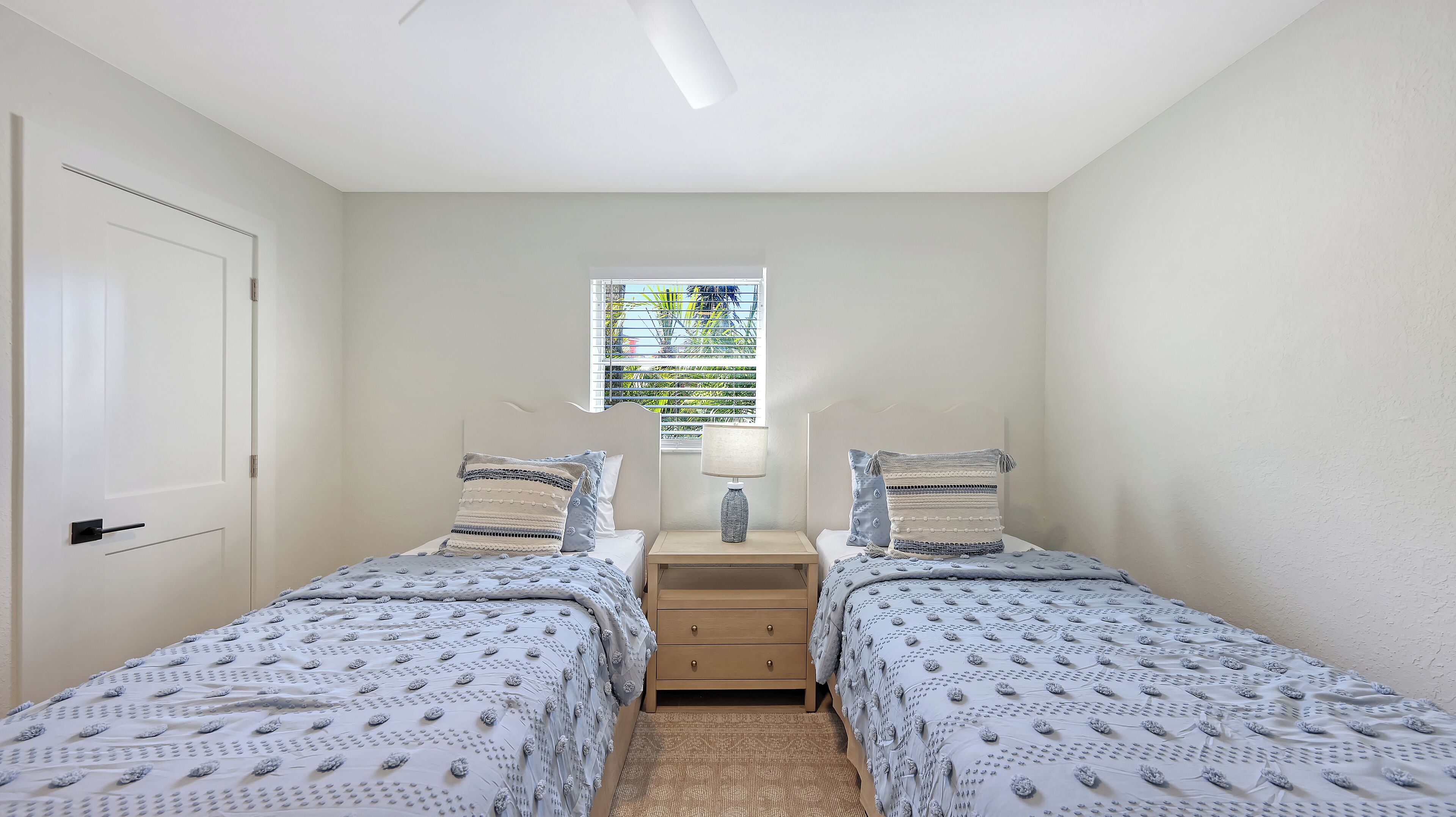A bedroom with two single beds, a nightstand with a lamp, and a window with blinds.