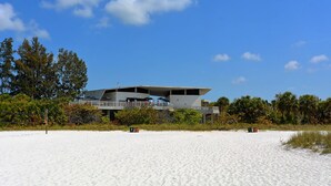 Un edificio moderno con techo plano y varios niveles, rodeado por una playa arenosa y abundante vegetación.