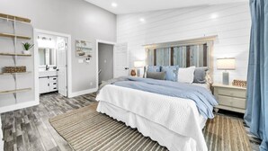 Un dormitorio moderno con una cama grande, ventilador de techo, cabecera de madera y un baño con tocador blanco y estantes.