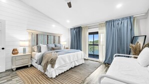 Un dormitorio con cama, mesitas de noche, ventilador de techo, vistas al exterior desde un balcón y un ventanal con cortinas.
