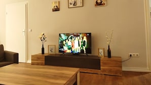 Ein Wohnzimmer mit einem Flachbildfernseher auf einem Holzboden, einer Couch und einem Couchtisch. An der Wand hängen drei gerahmte Bilder und auf dem Couchtisch steht eine Vase mit Blumen.