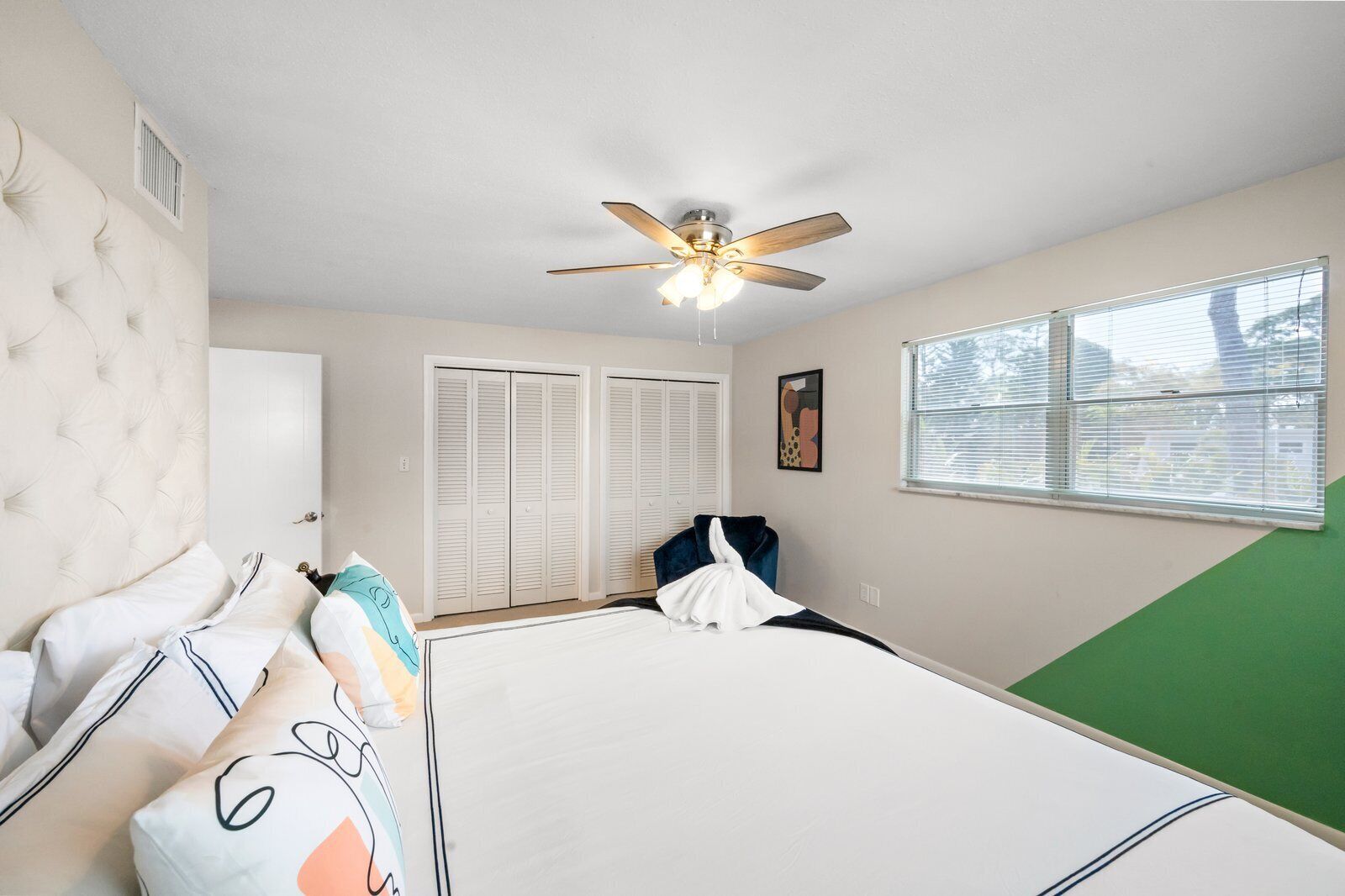 A bedroom with a tufted headboard, a bed with white bedding, a ceiling fan, a window with blinds, and a green accent wall.