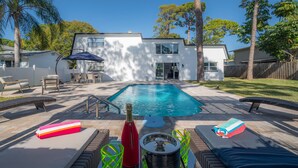 Un patio trasero con piscina, sillas de descanso, una mesa con una botella y vasos, y un antorcha tiki.