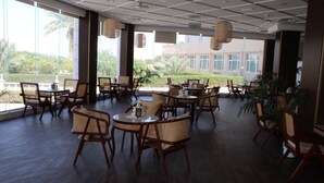 A spacious indoor dining area with multiple wooden tables and chairs, large windows offering a view of greenery and buildings outside. - Al Salam Grand Hotel & Resort (Al Buraymi)