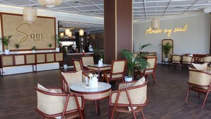 A café interior with round tables and wicker chairs, a reception desk, and a neon sign. - Al Salam Grand Hotel & Resort (Al Buraymi)