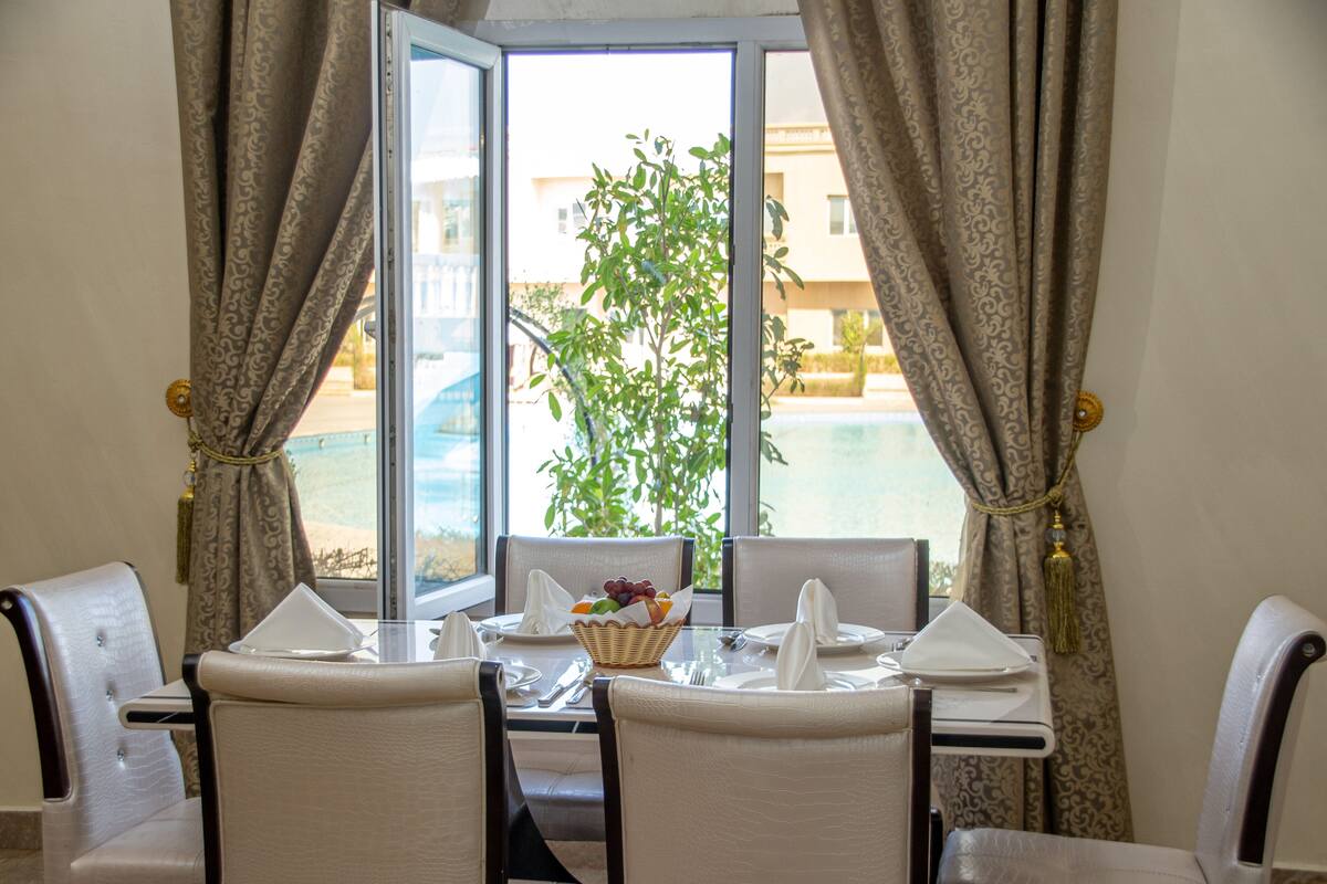 A dining area with a table set for four, a window with curtains, and a view of a building and pool outside.