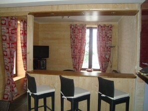 Una stanza con rivestimenti in legno, una zona bar, due sgabelli da bar, un televisore e tende a motivi decorativi.