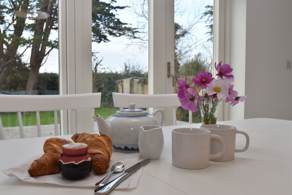 Desayuno con cruasanes, un cebrero, una jarrón y tazas sobre una mesa junto a una ventana con flores.
