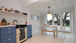 Een moderne keuken met een fornuis, oven en diverse keukengerei. Er staat een eettafel met stoelen en een bank. De keuken heeft witte kasten en een raam met uitzicht op een tuin.