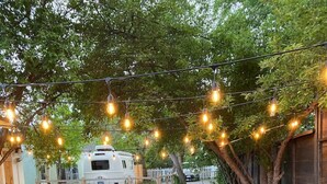 Un patio al aire libre con una mesa y sillas, luces colgantes y una caravana blanca al fondo.