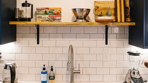 Una cocina con salpicadero blanco de azulejos, un lavamanos de acero inoxidable y una encimera de madera. Sobre el lavamanos, un cuadro enmarco con un paisaje.