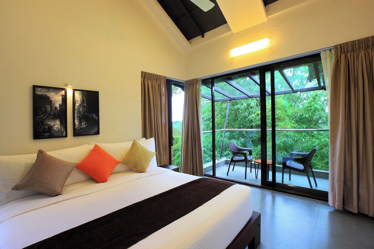 A hotel room with a large bed, two pillows, a blanket, a nightstand, a window with curtains, and a view of greenery outside.