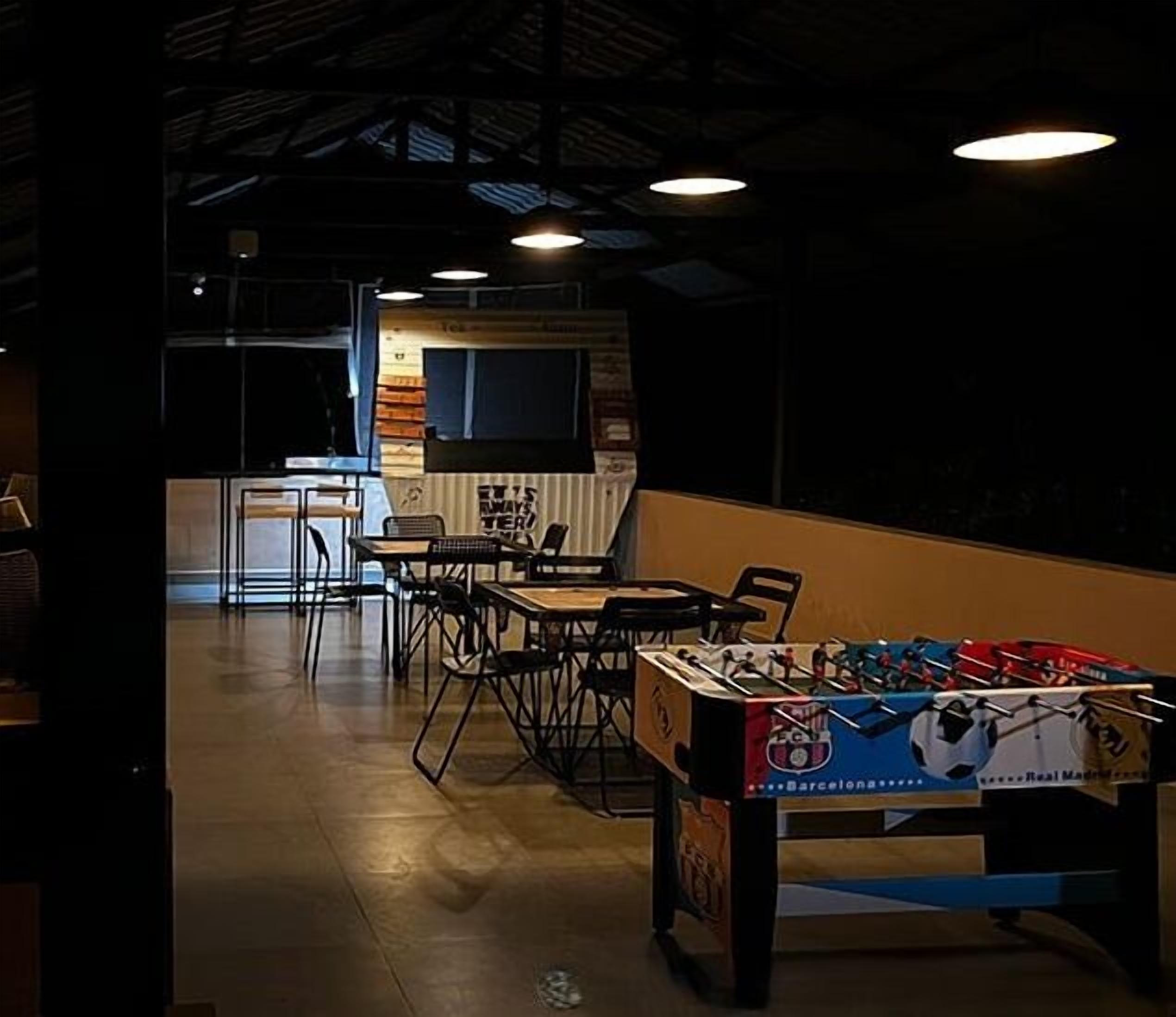 A dimly lit indoor space with a foosball table, tables, and chairs arranged for dining or socializing.