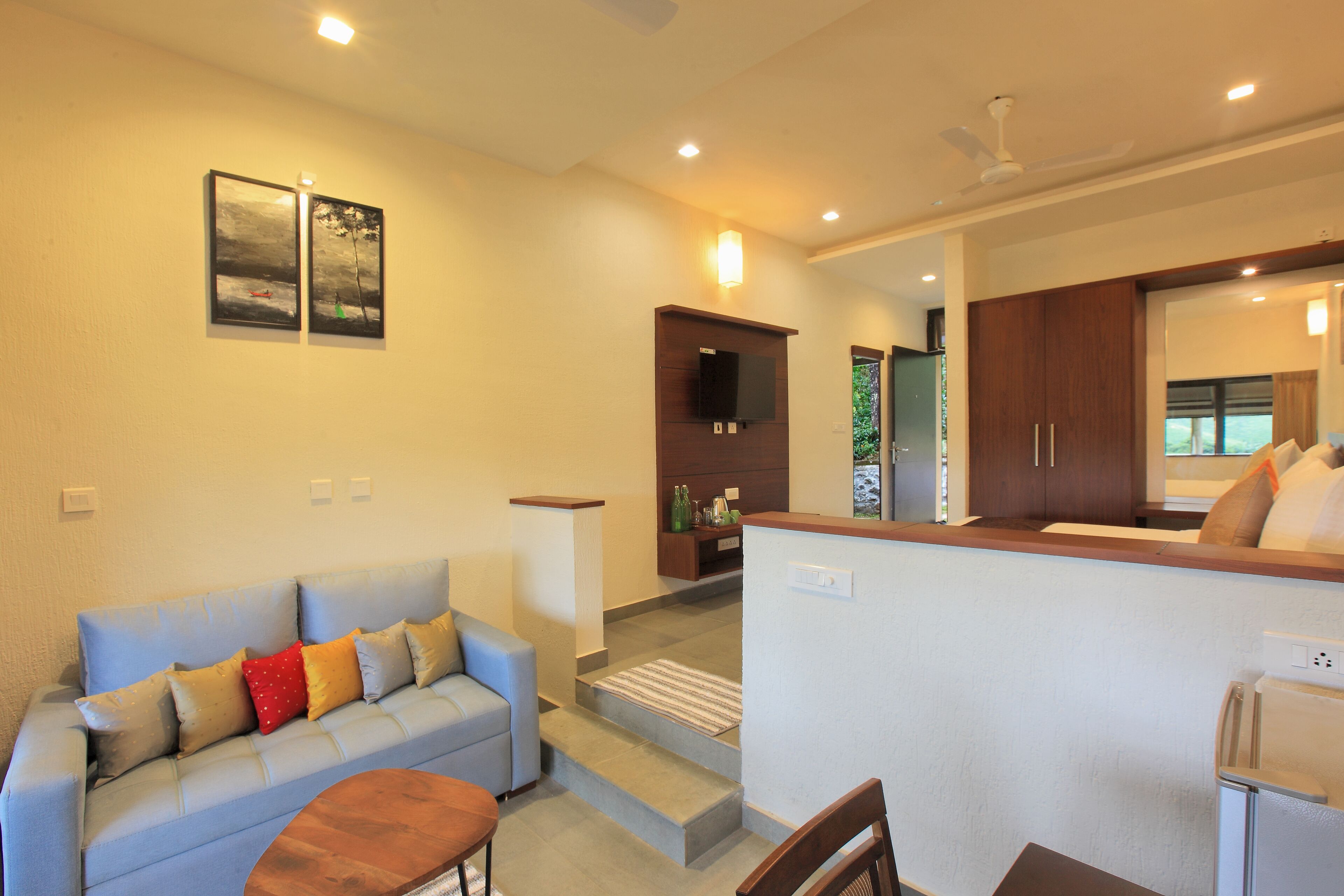 A modern living room with a sofa, a coffee table, and a TV mounted on the wall. There is a kitchen area visible in the background with wooden cabinets and a refrigerator.