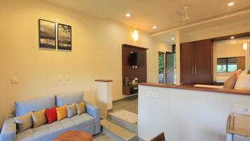 A modern living room with a sofa, a coffee table, and a TV mounted on the wall. There is a kitchen area visible in the background with wooden cabinets and a refrigerator.