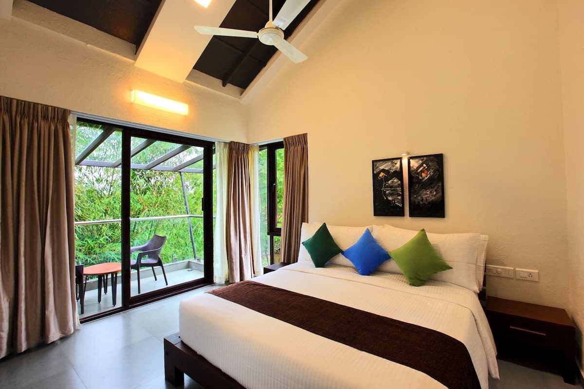 A bedroom with a large bed, a ceiling fan, a nightstand, and a view of greenery through the window.