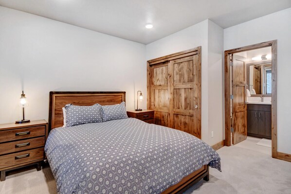 A bedroom with a wooden bed, two bedside tables with lamps, a wardrobe, and a bathroom visible through an open door.