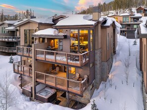 A modern multi-story lodge with a balcony and hot tub, surrounded by snow.