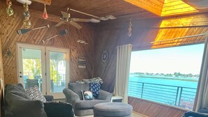 A cozy cabin interior with a wooden ceiling, a sofa, a round ottoman, a small table, and a view of the water through large windows. - Lake Front home w/Dock & Boat Launch ✨ Closer to the Sun Lake House (Moses Lake)