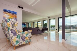 A spacious living area with a large armchair, sofas, and a glass wall offering a view of an outdoor pool.