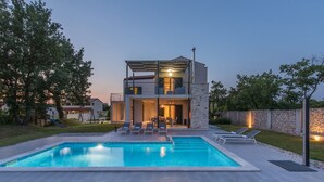 Una casa moderna de dos pisos con piscina, mobiliario exterior y un muro de piedra.