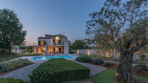 Una casa moderna de dos pisos con piscina, rodeada de jardines cuidados y un árbol grande.