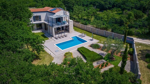 Una casa moderna de dos pisos con piscina, rodeada de exuberante vegetaci ón y un muro de piedra.