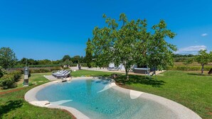 Une piscine dont le bord est recouvert de petits galets, entourée d’une pelouse et d’un arbre.