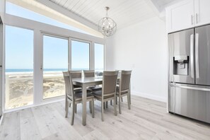 A modern kitchen with a large dining table and chairs, a stainless steel refrigerator, and a view of the beach through large windows.
