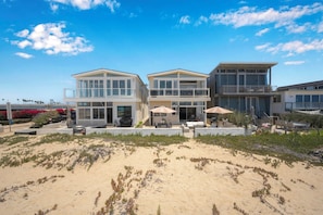 Een pand aan het strand met een tweelaags huis, grote ramen, zitplaatsen buiten en een zandstrand op de voorgrond.