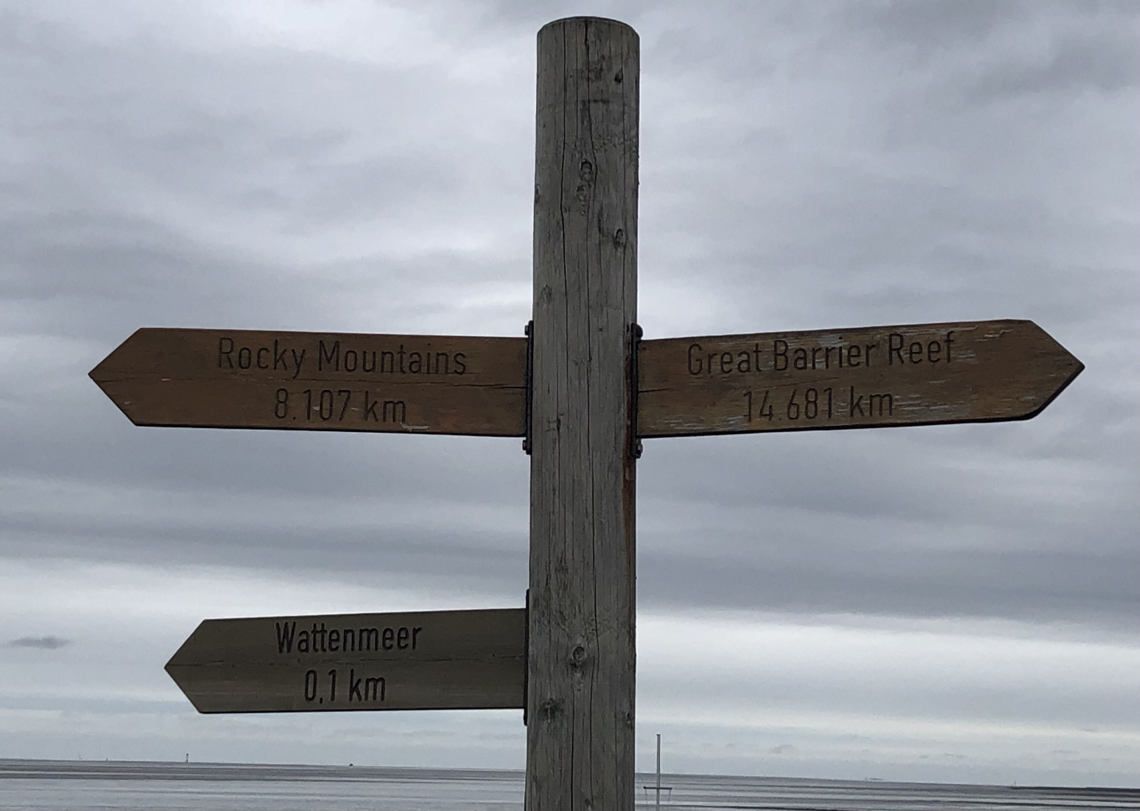 Ein hölzernes Wegweiser mit Pfeilen, die zu den Rocky Mountains, dem Great Barrier Reef und dem Wattenmeer zeigen, mit angegebener Entfernung.