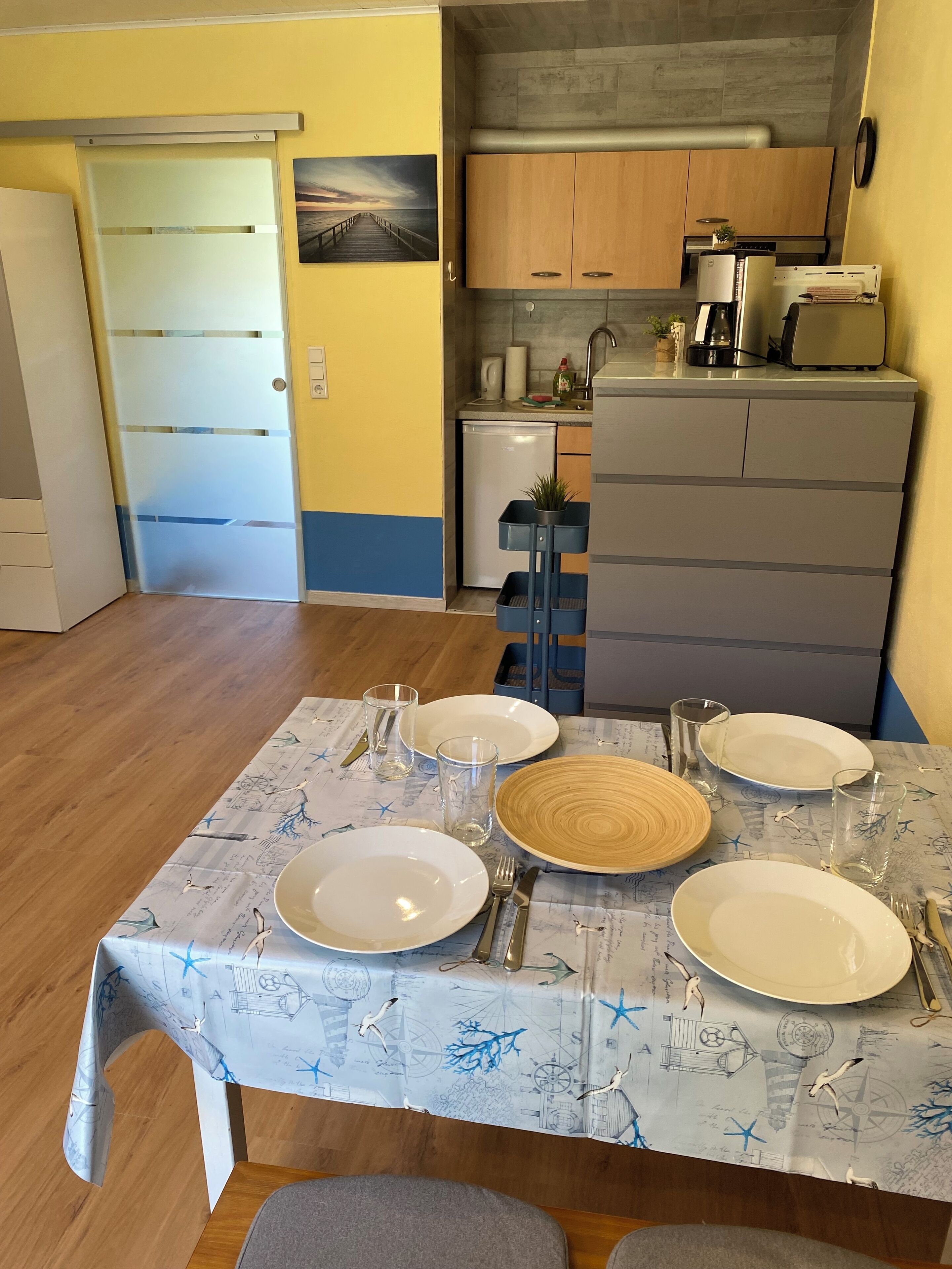 Ruokailutila, jossa on neljälle katettu ruokapöytä, taustalla keittiö ja ovi, joka johtaa toiseen huoneeseen.