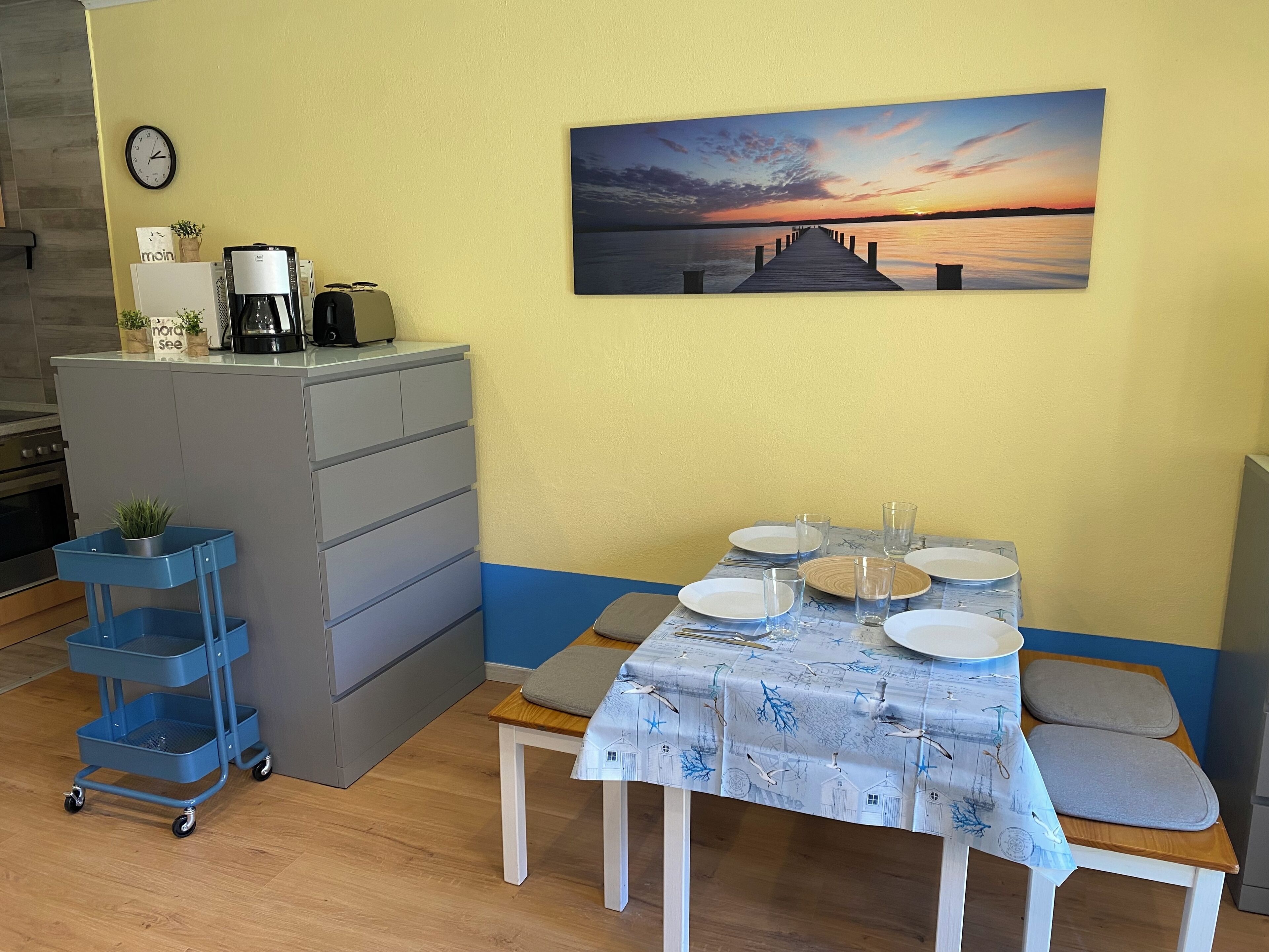 Ein Essbereich mit einem Tisch für vier Personen, einer Uhr an der Wand und einem Gemälde, das einen Sonnenuntergang über dem Wasser zeigt.