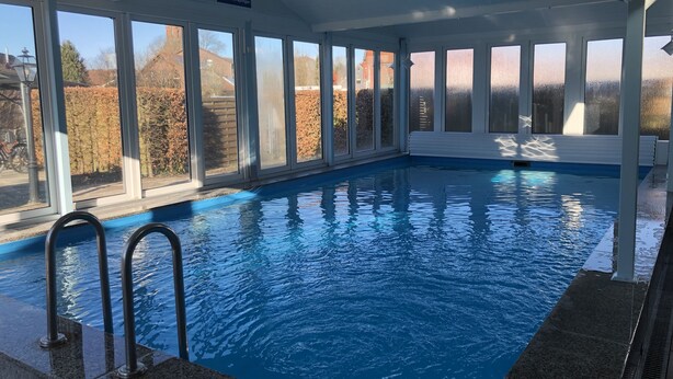 Une piscine intérieure avec des parois en verre transparent, un panneau indiquant qu’elle est réservée à un usage privé, et une vue sur un quartier résidentiel à l’extérieur.