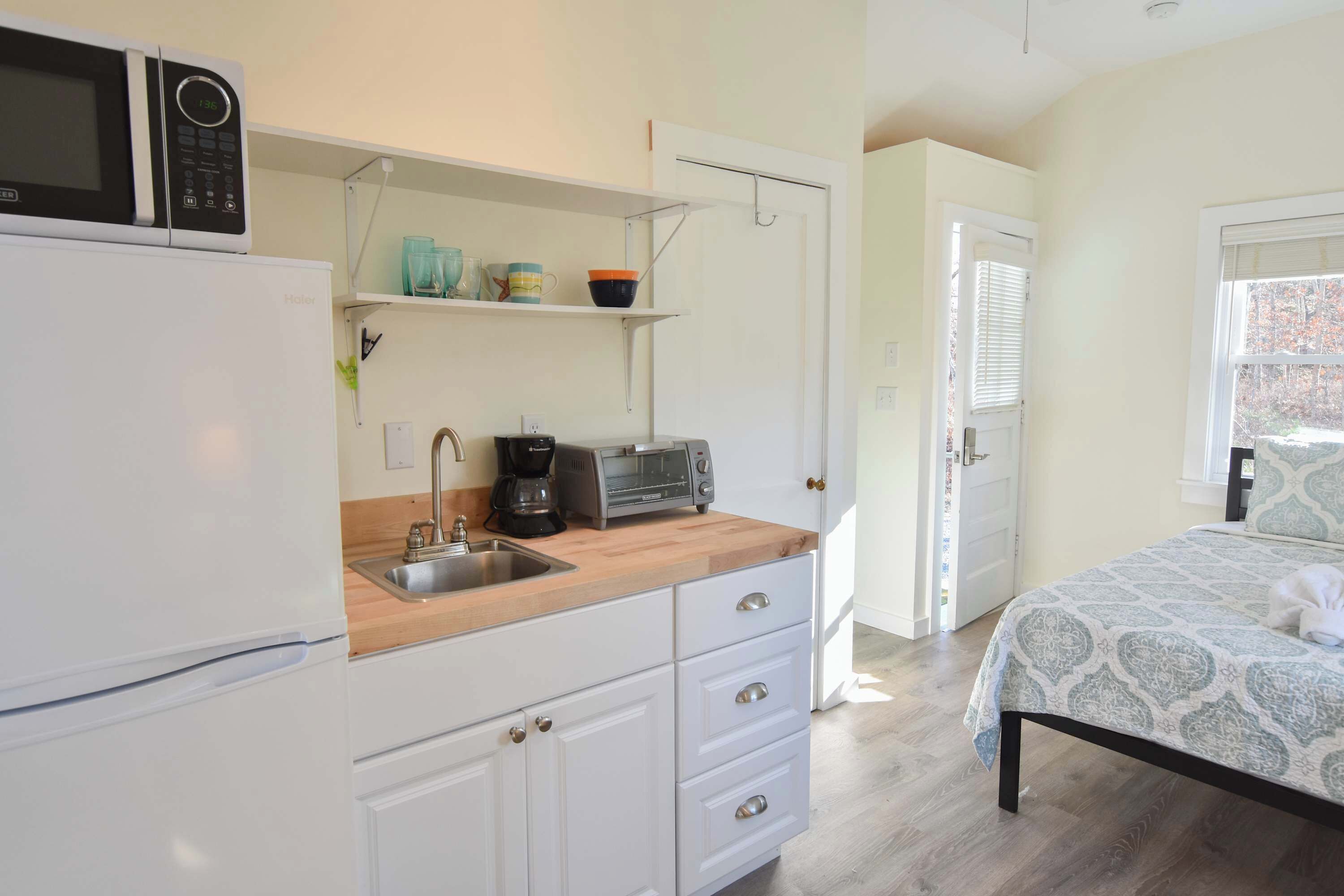 A compact kitchen with a microwave, coffee maker, and toaster on a wooden countertop. There is a white refrigerator, a sink, and a door leading to another room.