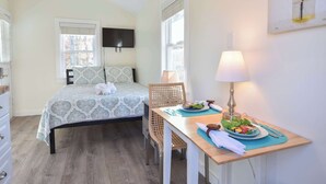 A bedroom with a bed, bedside table, lamp, and a dining area with a table, chairs, and a plate of food. - Cozy Colorful Cottage Perfect for 2! (Eastham)