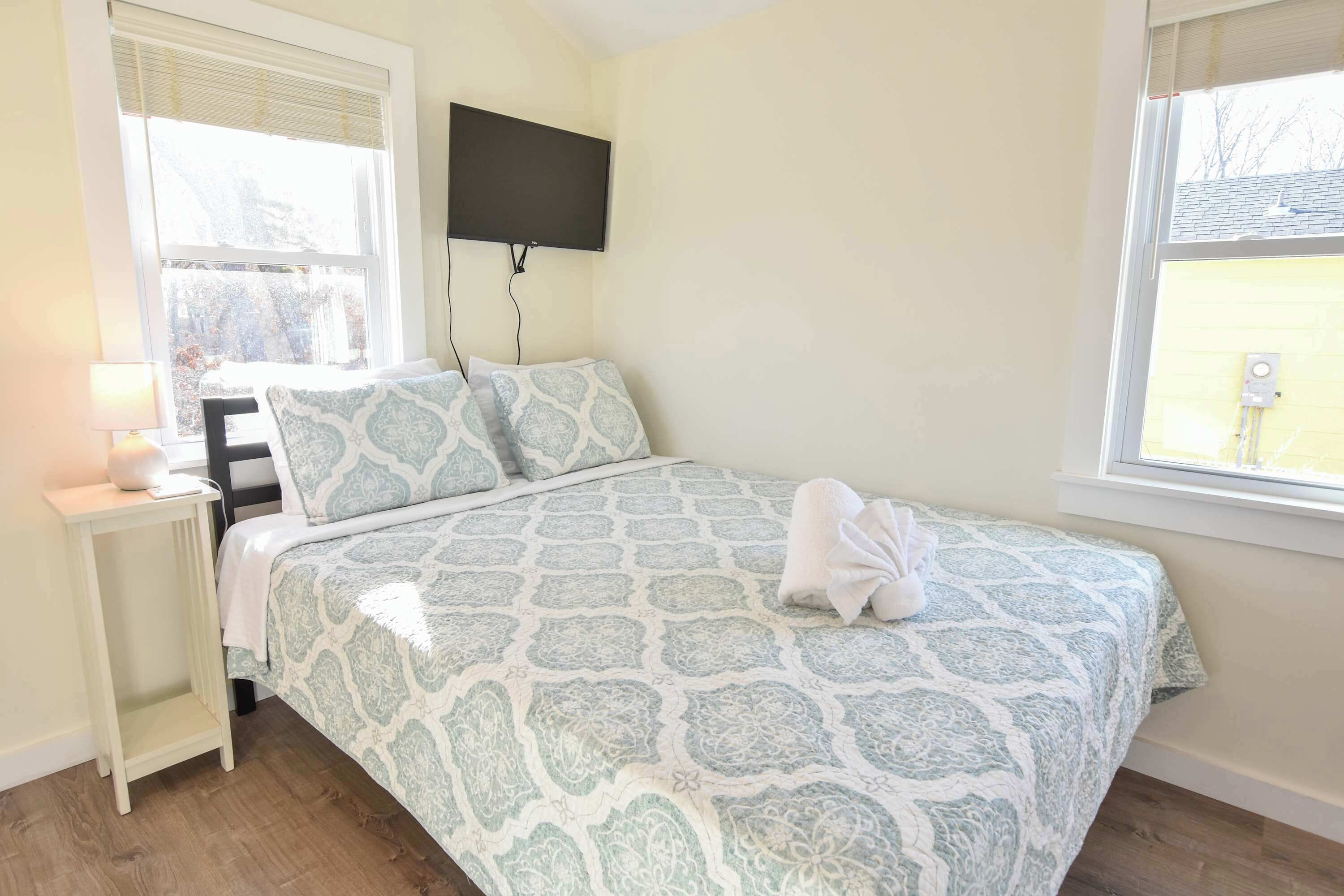 A bedroom with a bed, a bedside table with a lamp, a TV mounted on the wall, and a window with blinds.