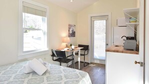 A compact room with a bed, a desk with a chair, a window, and a door leading to a kitchen area. - Dreamy Cottage for Two! Close to Beaches! (Eastham)