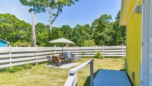 A backyard with a wooden deck, a white fence, and a yellow building. - Bright Cottage Close to Bayside Beaches! (Eastham)