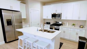 A modern kitchen with white cabinets, a stainless steel refrigerator, a microwave, a stove, and a sink. There are three white bar stools at the island. - Escondido Luxury Resort, pool, slide, pickleball, 4 bedrooms, bunks, 2car garage (Washington)