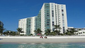 Ein hohes, mehrstöckiges Hotel mit Balkonen in Strandnähe, mit Palmen und einem Sandstrand.